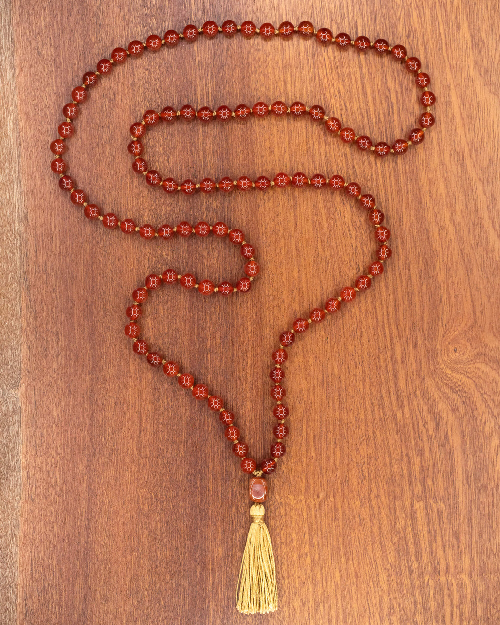 Japa mala de Cornalina completo, cuentas rojas en forma de S con borla dorada sobre madera oscura