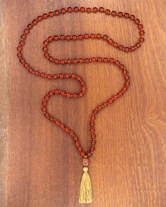 Japa mala de Cornalina completo, cuentas rojas en forma de S con borla dorada sobre madera oscura