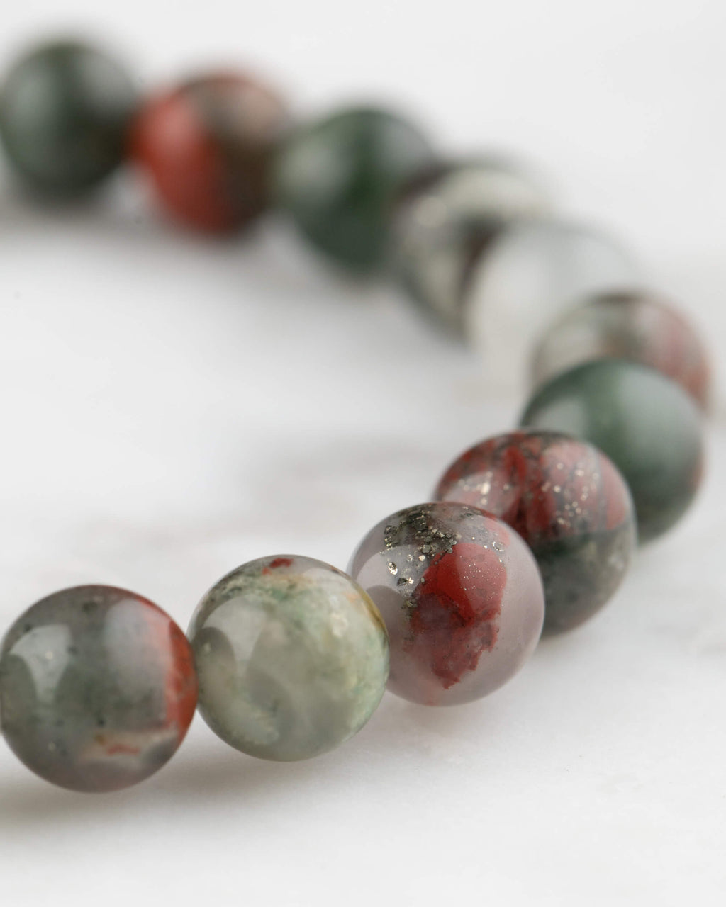 Pulsera de Piedra Sangre vista en angulo, cuentas verdes, grises y rojas sobre mármol claro