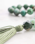 Detalle macro del nudo de la borla verde junto a una cuenta facetada gris oscura y cuentas verdes de fondo