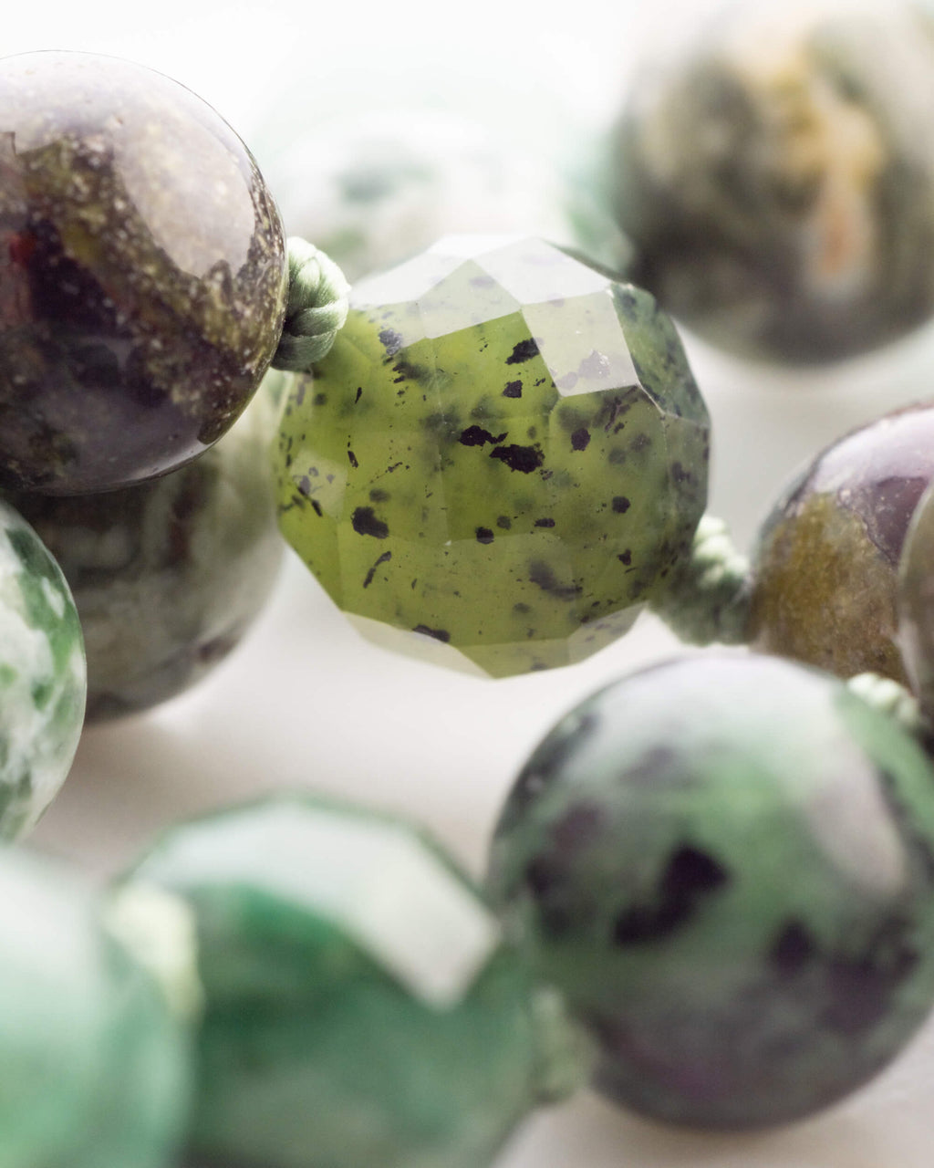 Detalle macro de una cuenta facetada verde oliva con manchas negras, junto a cuentas moradas y verdes moteadas