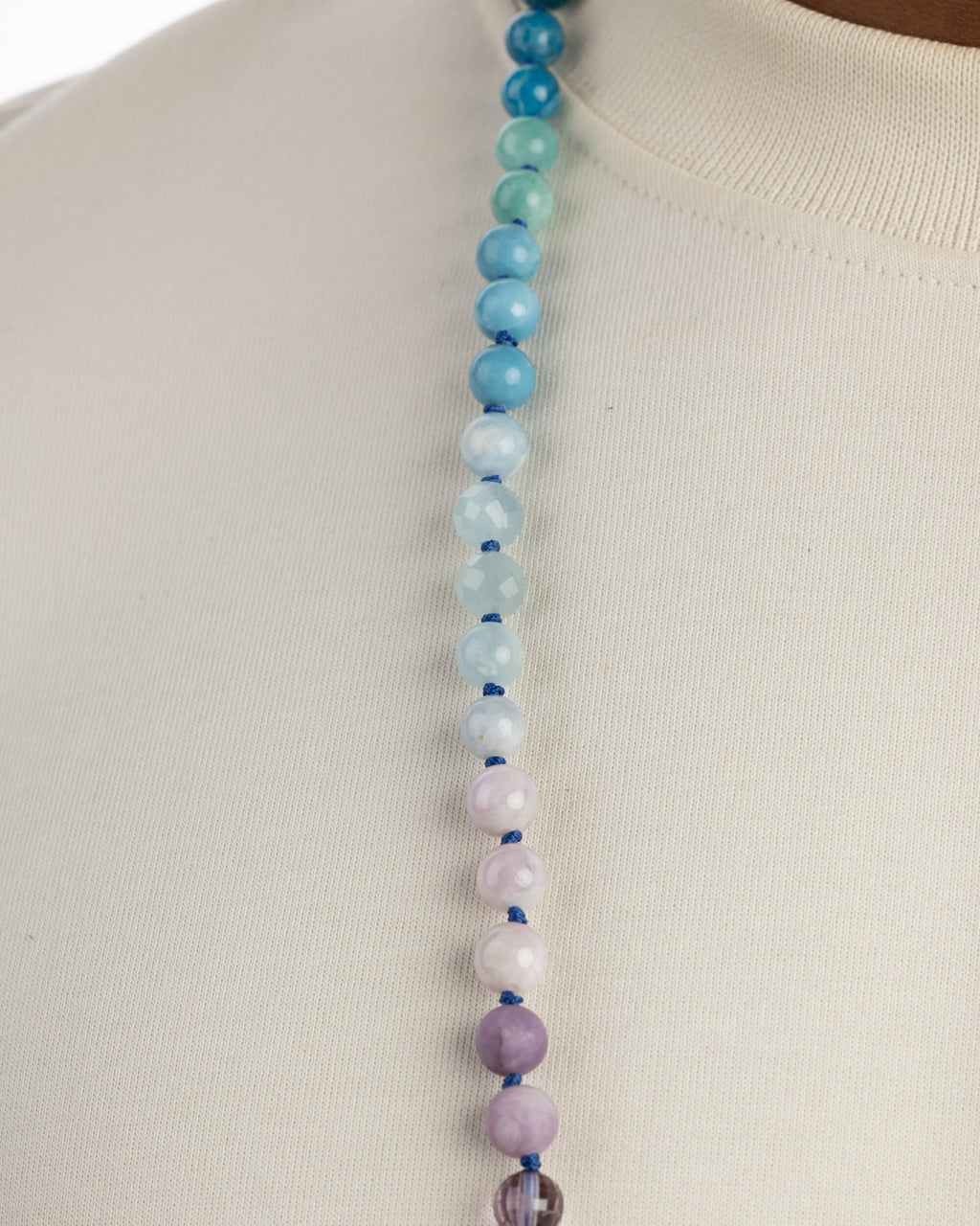Detalle macro de un collar con cuentas azules y verdes facetadas arriba y cuentas lila pastel opacas hacia abajo