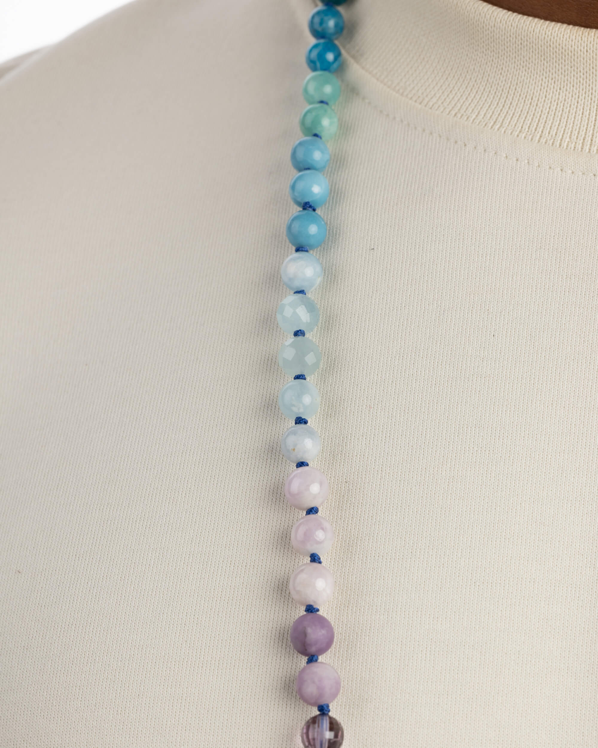 Detalle macro de un collar con cuentas azules y verdes facetadas arriba y cuentas lila pastel opacas hacia abajo